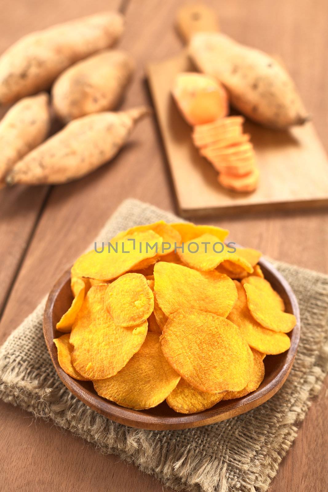 Crispy Peruvian sweet potato chips on wooden plate with sweet potatoes in the back (Selective Focus, Focus one third into the sweet potato chips)