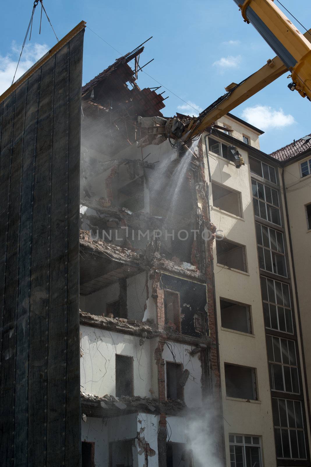 A large tracked demolition excavator fitted with concrete crunching demolition tool, ripping apart an old multi storeyed building whilst a heavy duty crane is applying a rubber mat to protect the surroundings