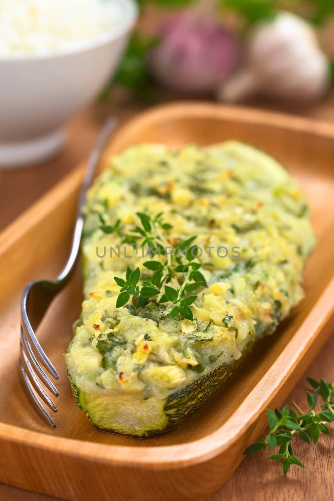 Baked zucchini stuffed with mashed potato, cheese and herbs (thyme, oregano, parsley, garlic) garnished with thyme served on wooden plate with a fork beside and a bowl of cooked rice in the back (Selective Focus, Focus on the front of the thyme garnish on the zucchini)