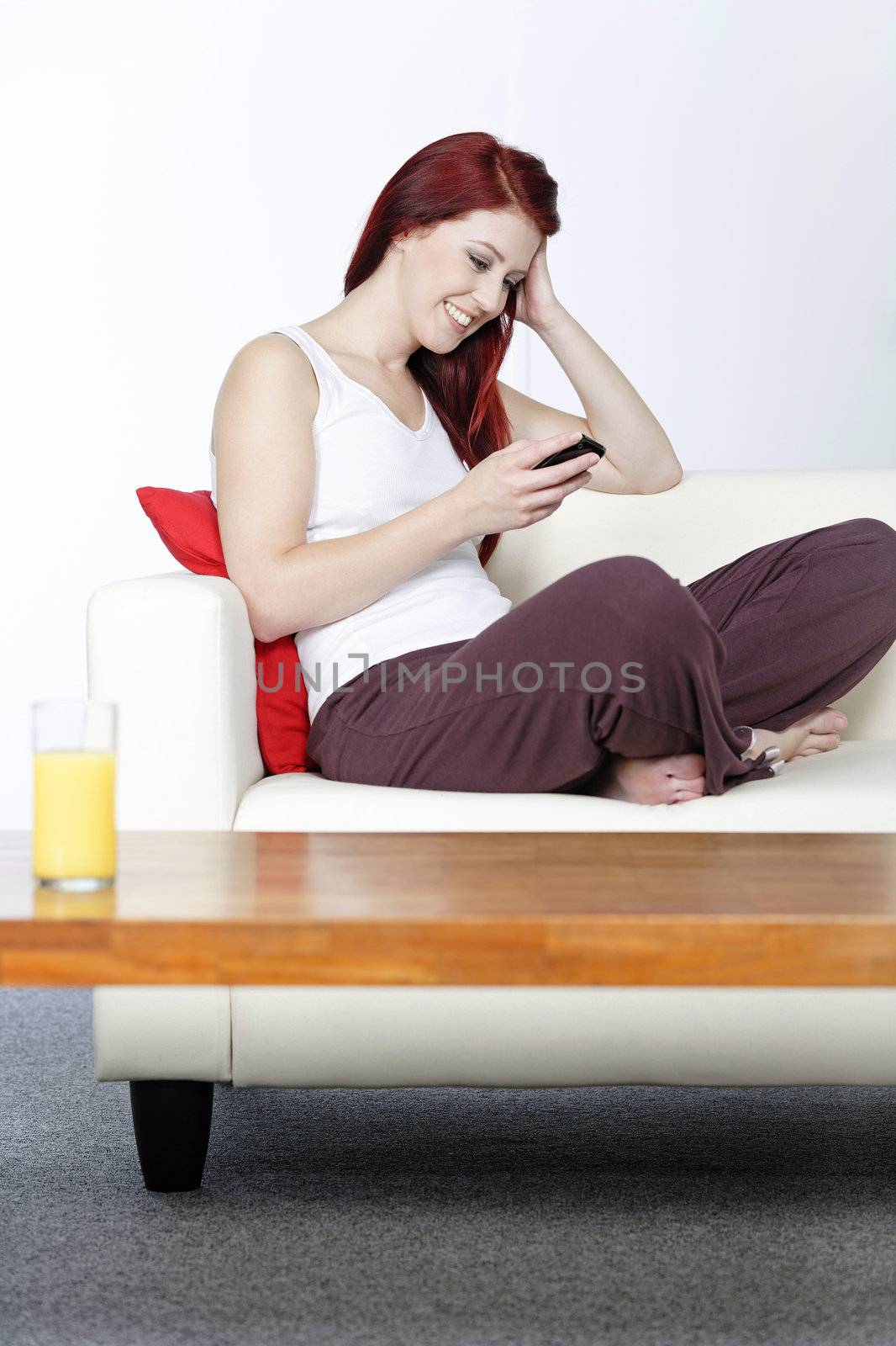 happy young woman sat on her white sofa at home smiling at text messages on her phone