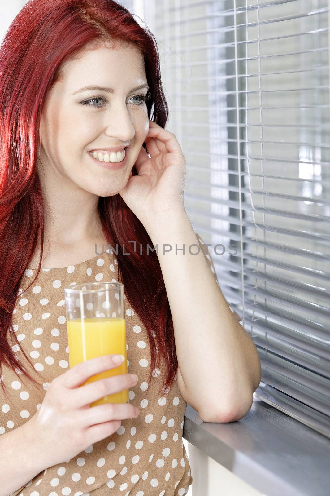 Happy smiling young woman standing by a window at home with a fresh glass of Orange juice.