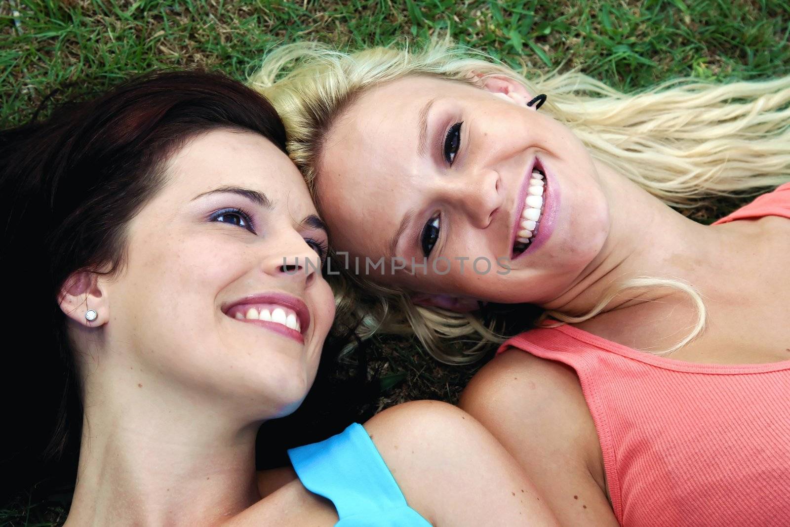 Two gorgeous smiling young women friends lying on green grass and looking up