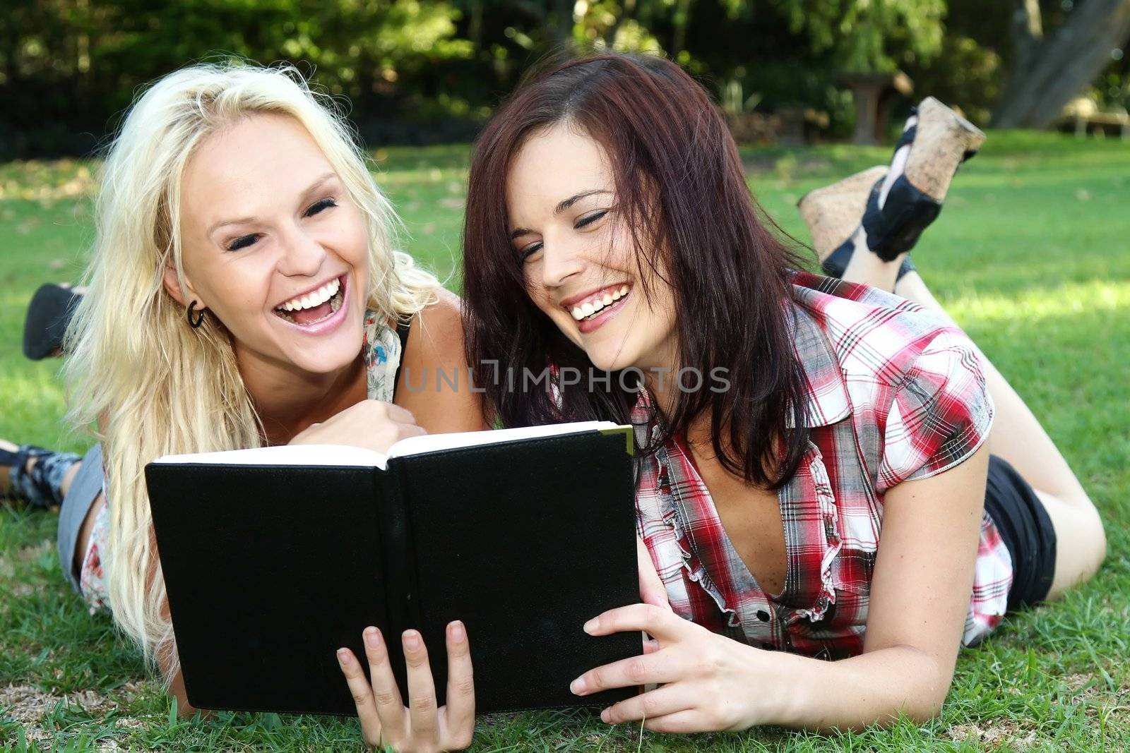 Lovely laughing brunette and blonde girls reading a book while lying on the green grass outdoors