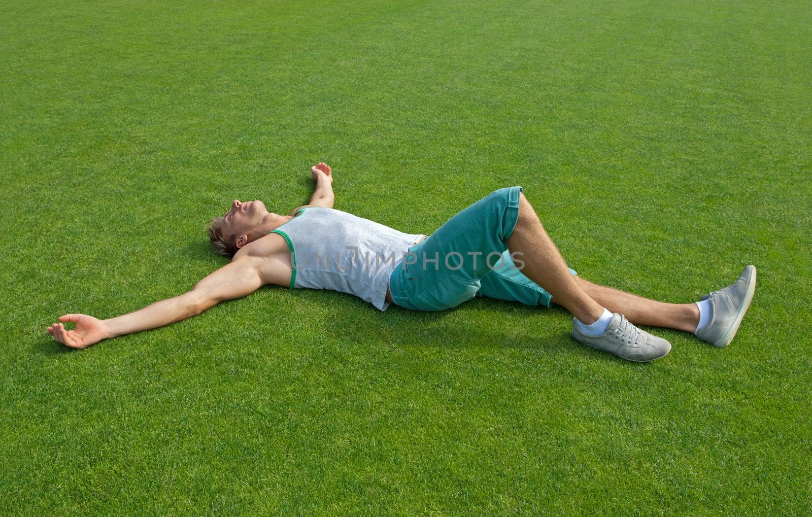 Sporty young man laying on green training field with his arms spread, relaxing.