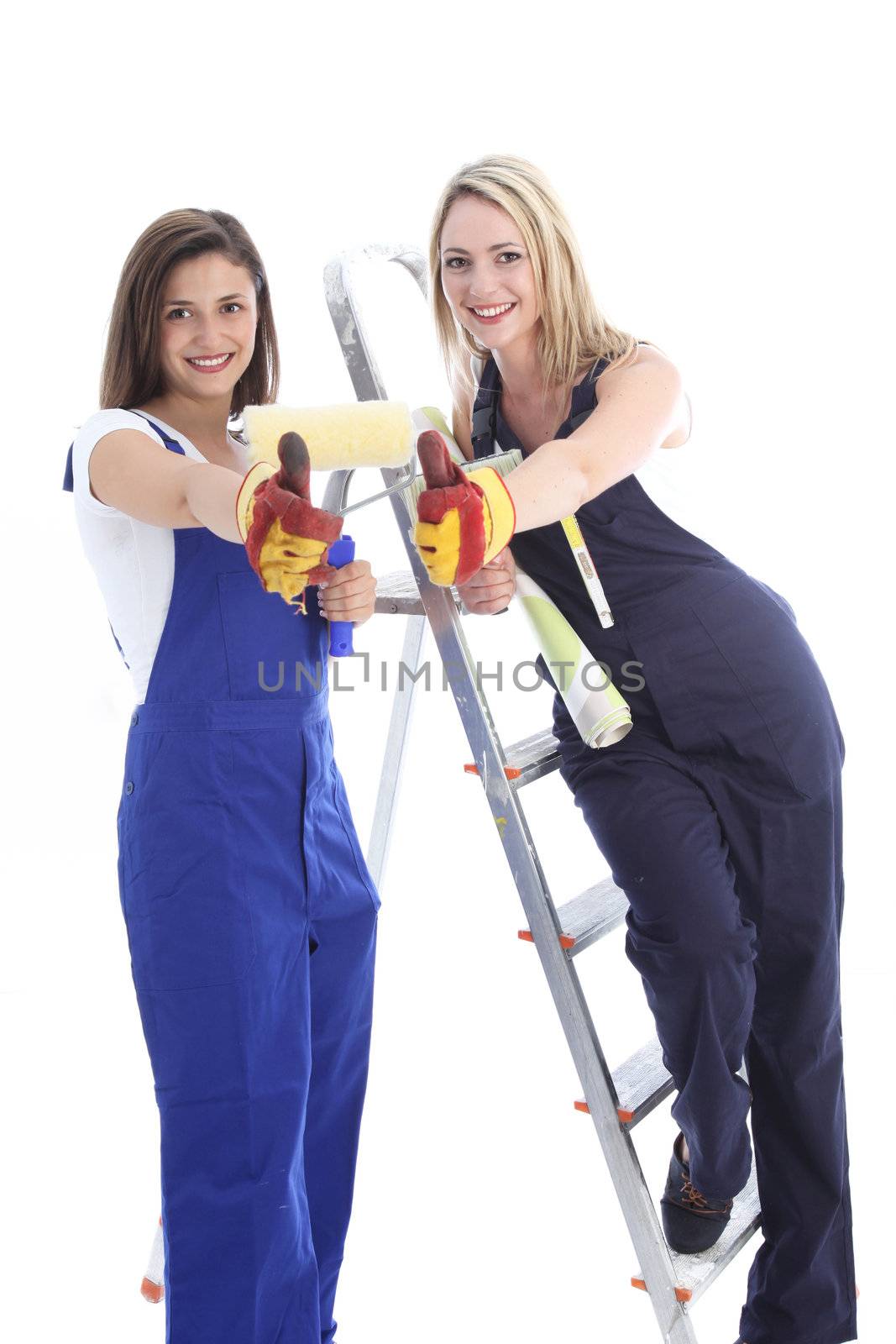 Two happy women dressed in dungarees and safety gloves give a thumbs up for the benefits of teamwork in hanging wallpaper and redecorating 