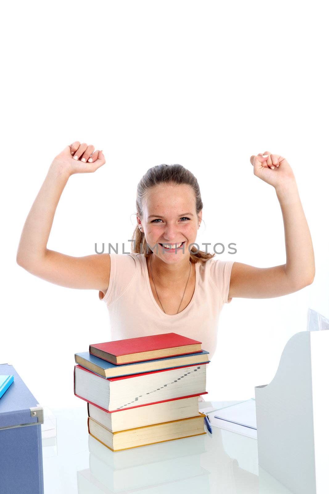 Jubilant young student sitting at her desk in front of a pile of textbooks with her hands raised in the air in celebration 