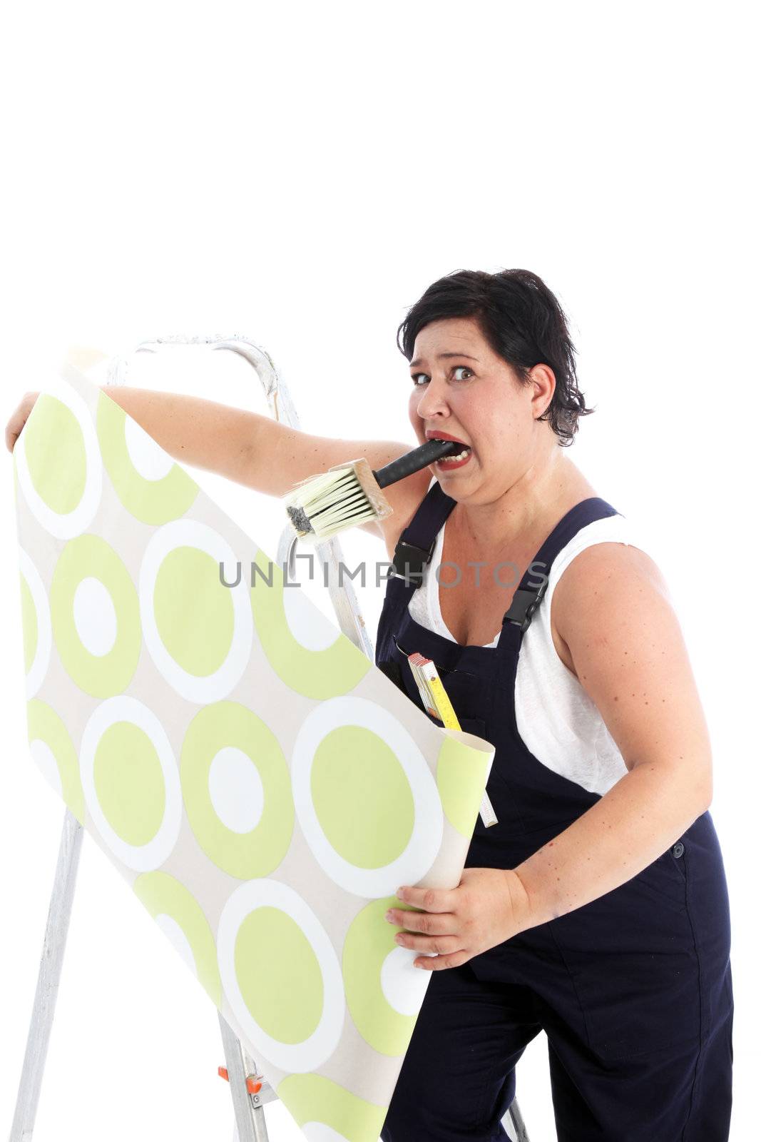 Exasperated woman hanging wallpaper with a brush full of glue paste clenched in her mouth as she holds the roll of paper in both hands Exasperated woman hanging wallpaper with a brush fulll of glue paste clenched in her mouth as she holds the roll of paper in both hands 