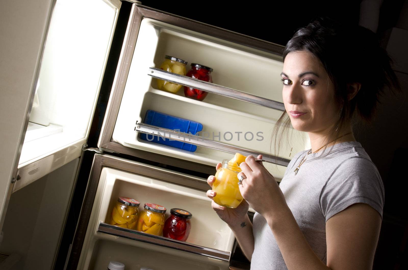 A sleepy woman lingers at the refrigerator door and gets suprised when you catch her nibbling outside her diet plan