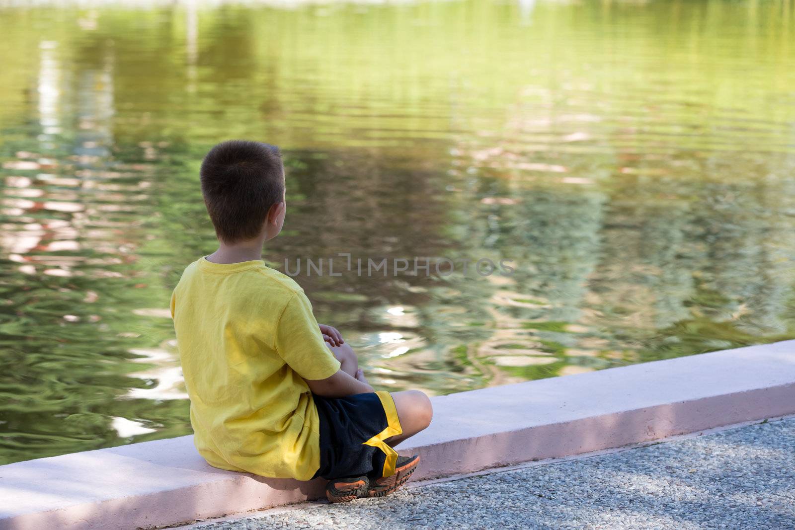 He is alone or lonely, seven years old kid looking at calm water while in deep thoughts.