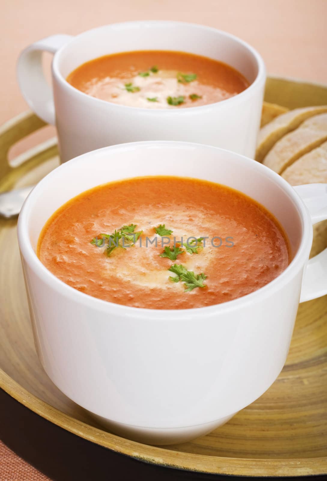 Two mugs of tomato soup with a swirl of cream and parsley, on a tray with bread.