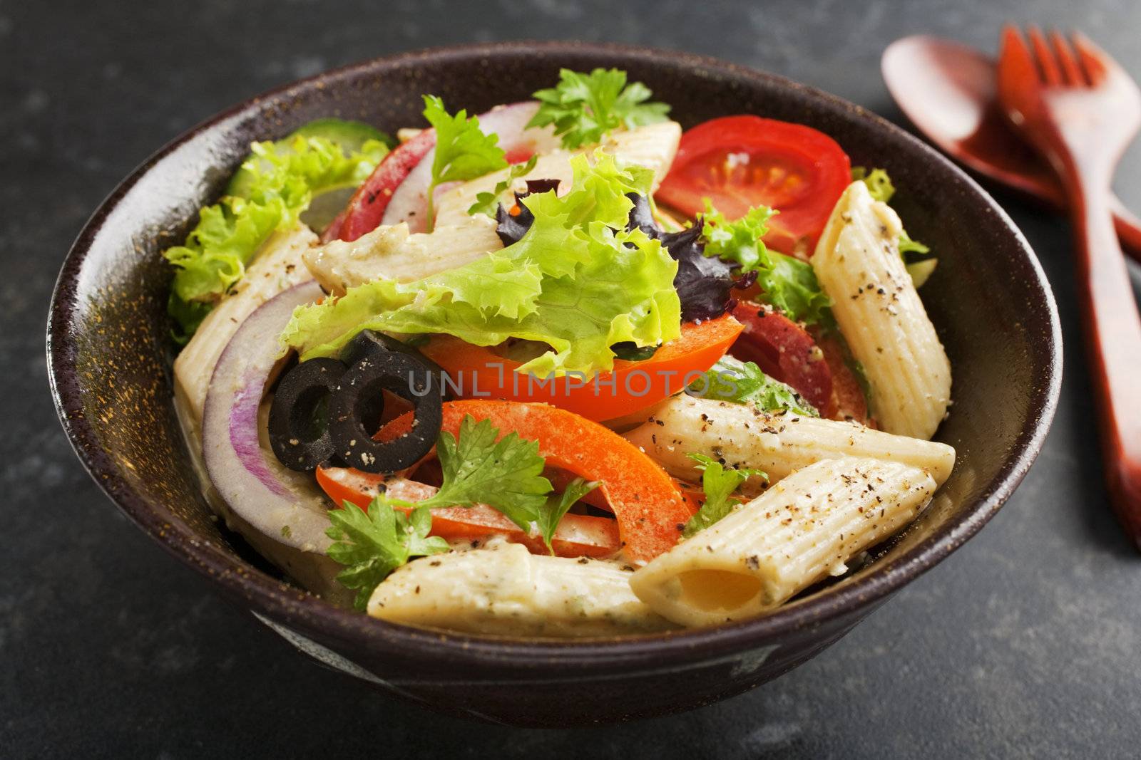 A bowl of pasta salad with a creamy pesto dressing. Salad includes red onions, curly lettuce, black olives, tomatoes,  bell peppers.
