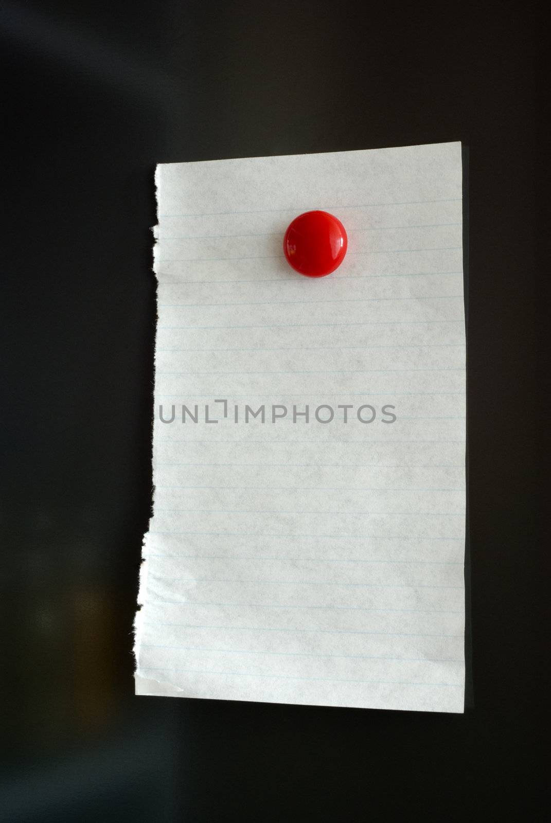 Piece of lined paper attached to fridge with a red button magnet. Fridge is dark and some parts of kitchen are slightly reflected.