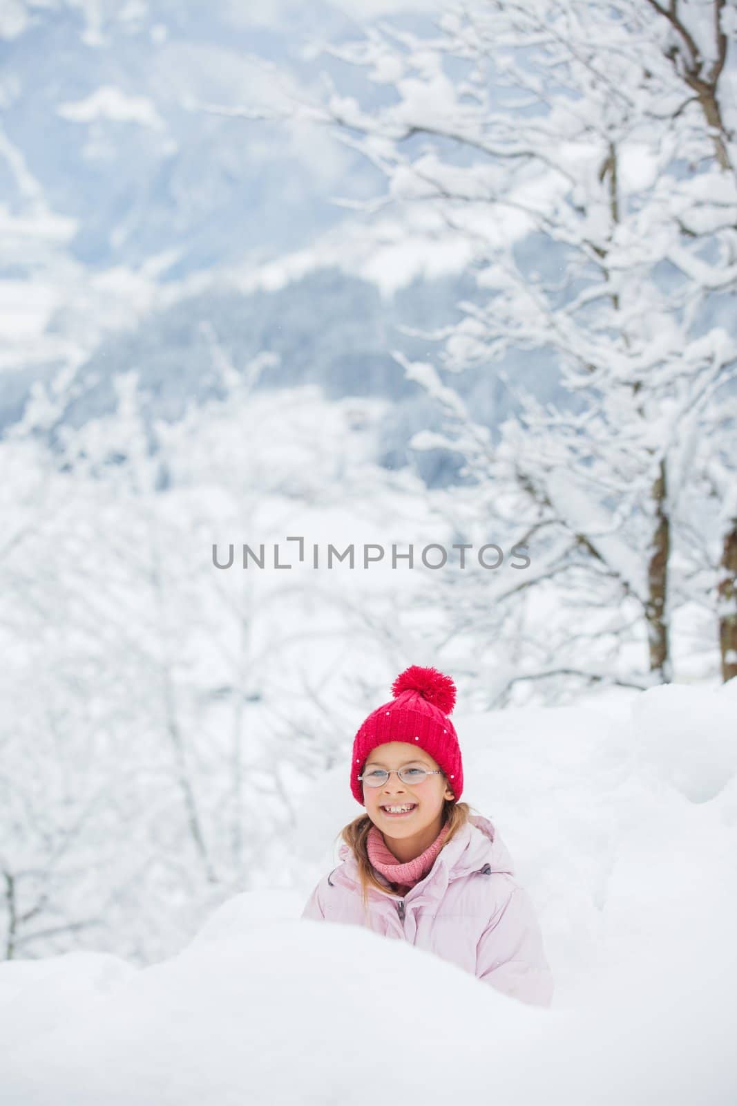 Happy girl in red hat smiling and having fun outdoors on snowing winter day, making snowball and loking at the camera