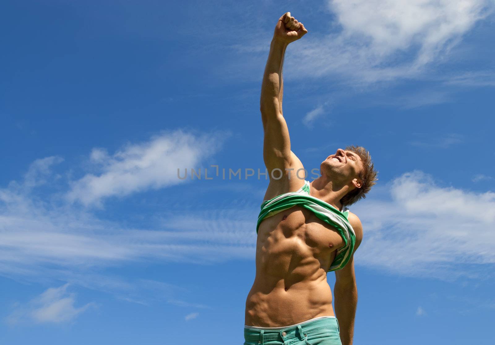 Happy fit man with his arm raised in joy, on blue sky background.