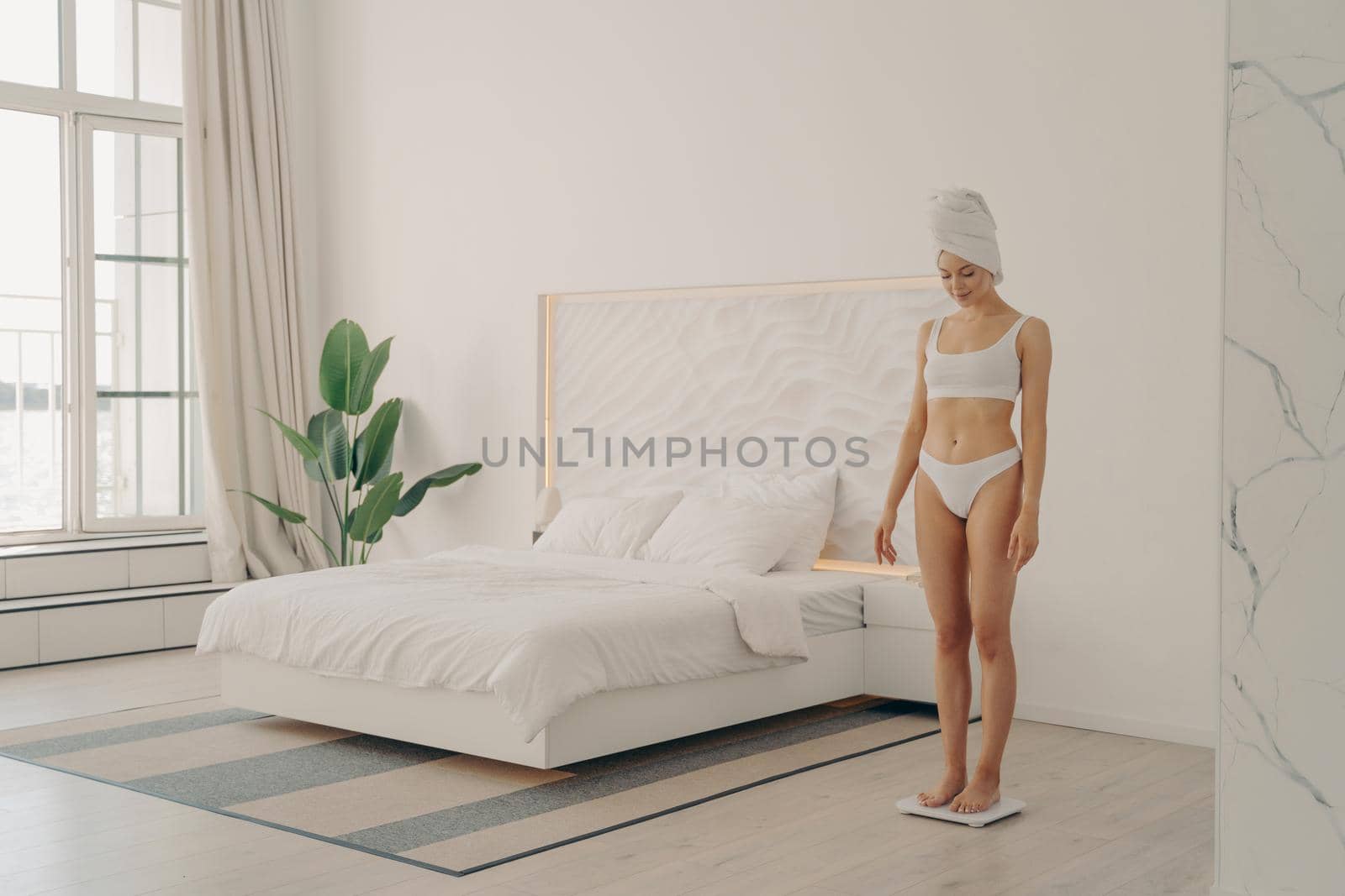 Shot of healthy young slender woman in underwear standing on scales in bedroom by vkstock