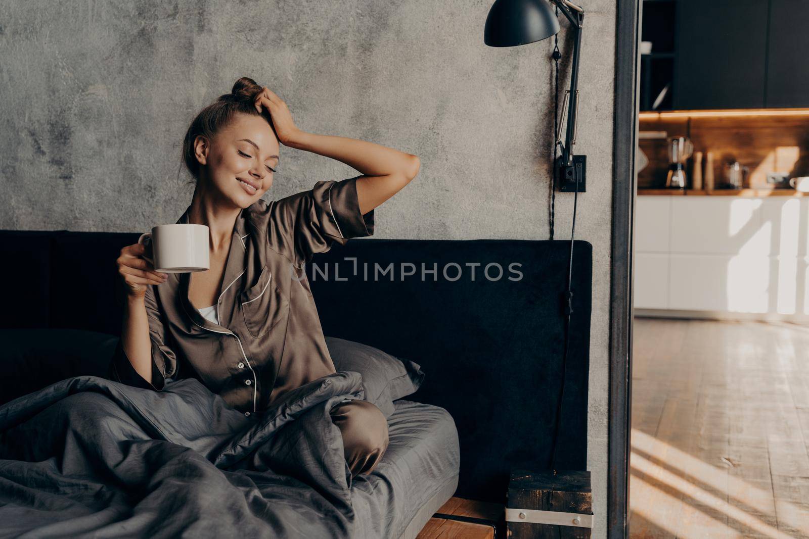 Portrait of young relaxed woman in stylish home suit waking up with cup of coffee in bed by vkstock