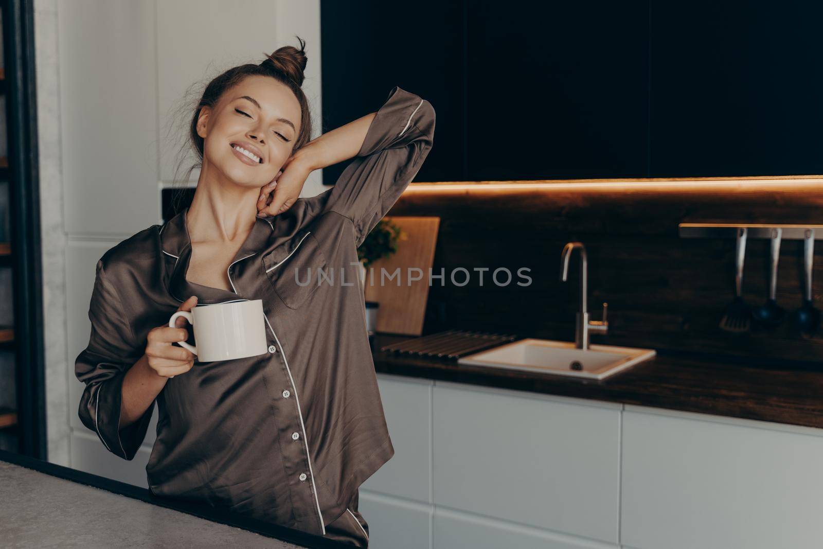 Beautiful relaxed young female in cozy silk pajama stretching from night sleep waking up early in morning with cup of coffee in her hand while standing in stylish kitchen interior at home