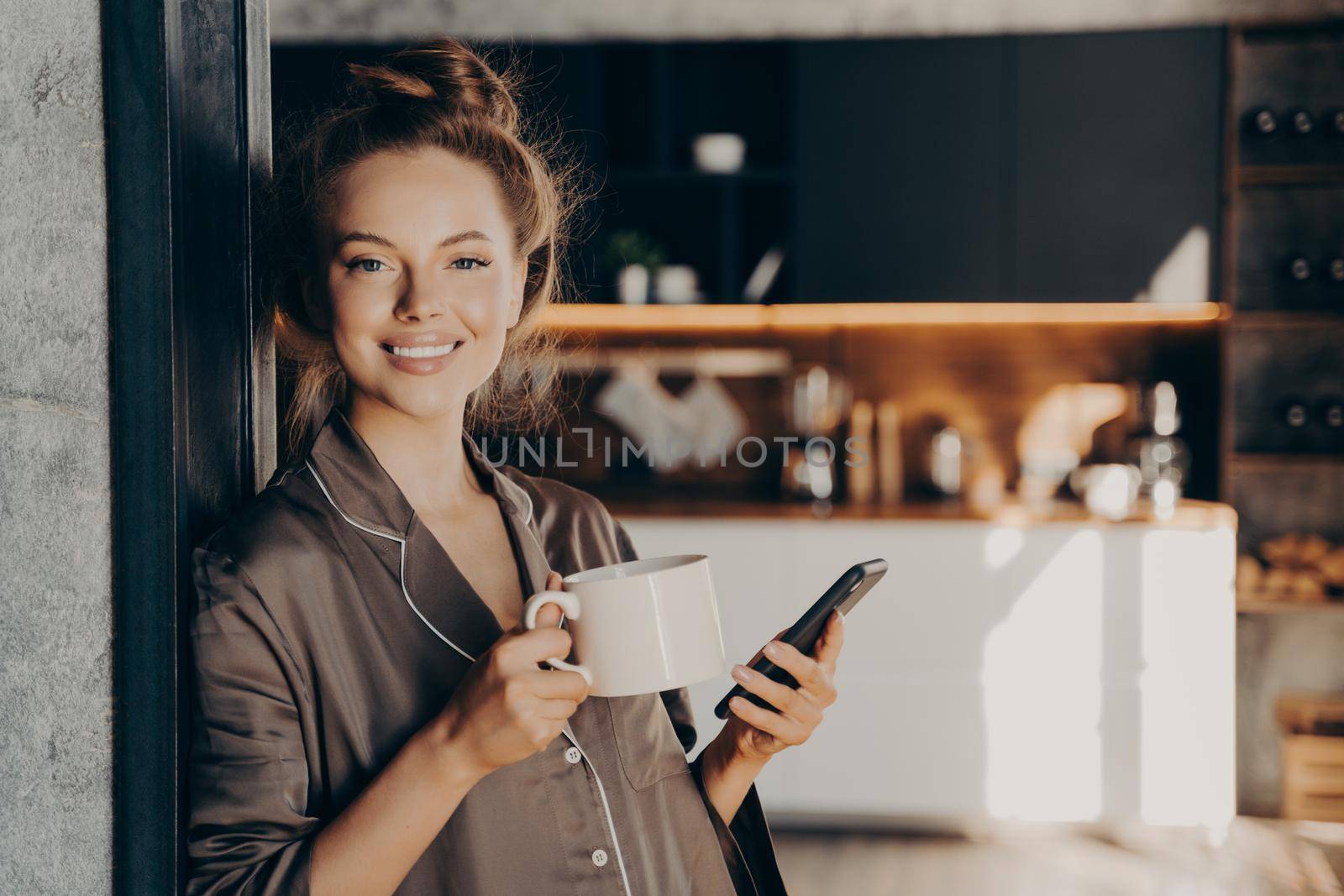 Lovely happy young woman having her morning coffee while checking new emails and notifications on smartphone standing in kitchen broadly smiling after waking up, spending weekend at home