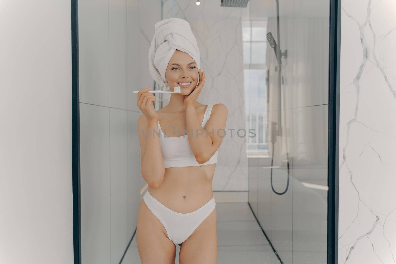 Young female model with fit body in white classic underwear standing in bathroom after taking shower while brushing her teeth. Personal body care and oral hygiene concept