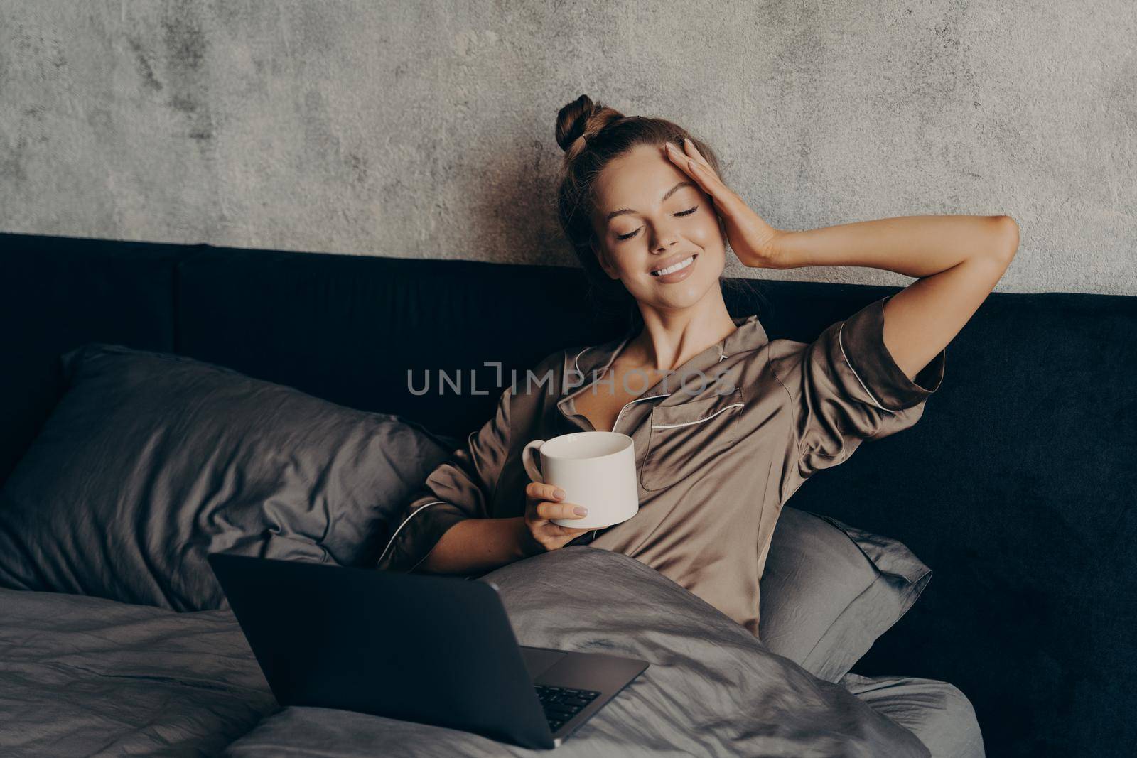 Charming relaxed young woman lying in bed at home with laptop and having morning cup of coffee, watching her favorite TV show online on computer, spending leisure time at home on weekend