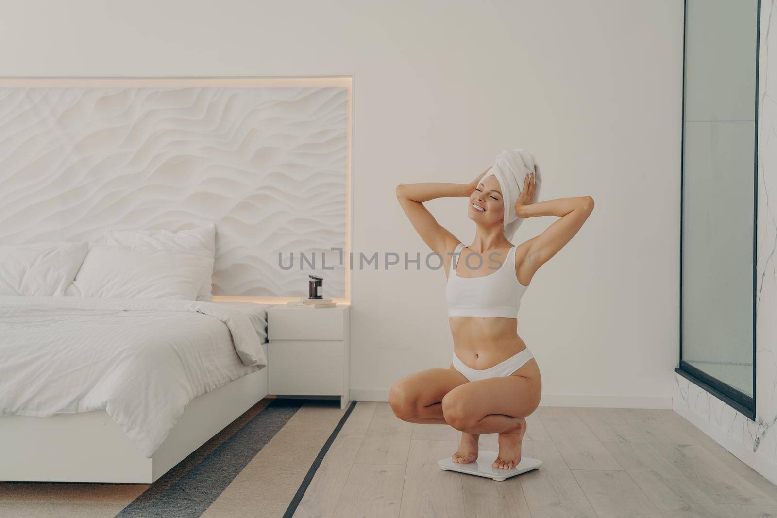 Beautiful young fit female model squats on scales in modern bedroom interior after morning shower routine in underwear happy and satisfied with result of her weight. Weight loss and dieting concept