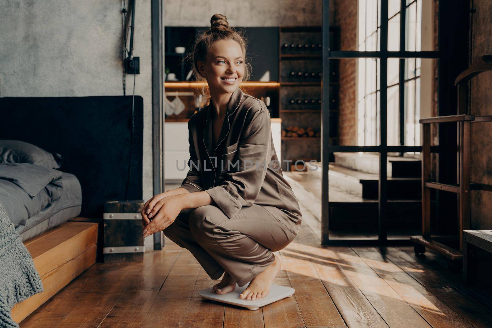 Image of joyful young sitisfied model in satin cozy pajama squatted over scales with wide smile in modern bedroom interior in morning, happy to see result of her dieting efforts, weight loss concept