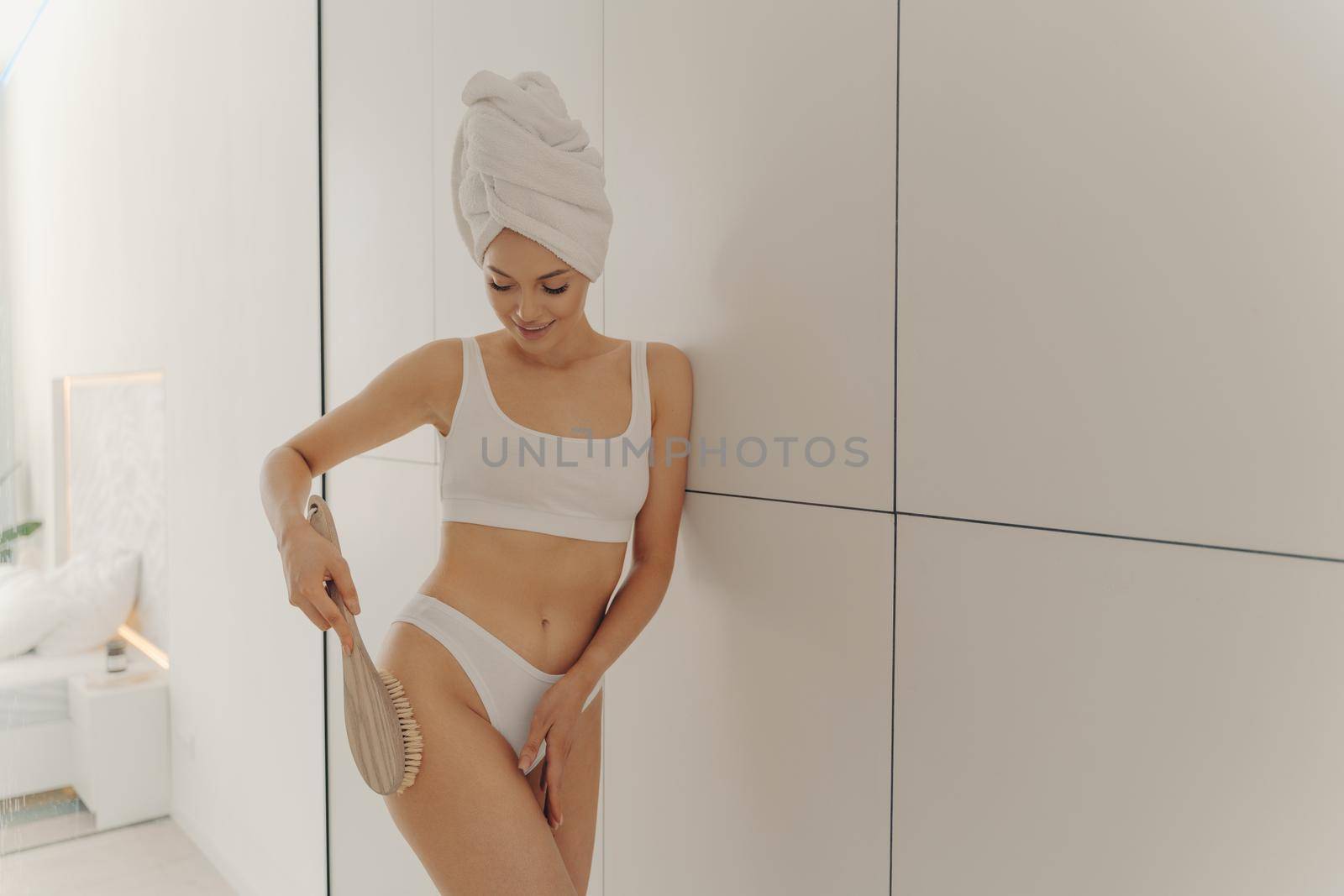 Beautiful young woman standing in white colored bathroom while making lymphatic drainage massage of legs with dry wooden brush holding it by long handle. Body care concept at home interior