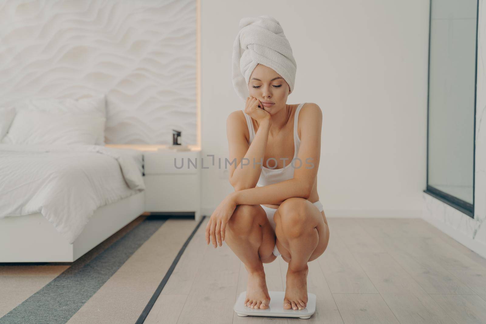 Unhappy young caucasian female squatting on tiptoes over electronic scales in bedroom with sad expression on her lovely face, not happy to see weight gain. Dieting and weight loss concept