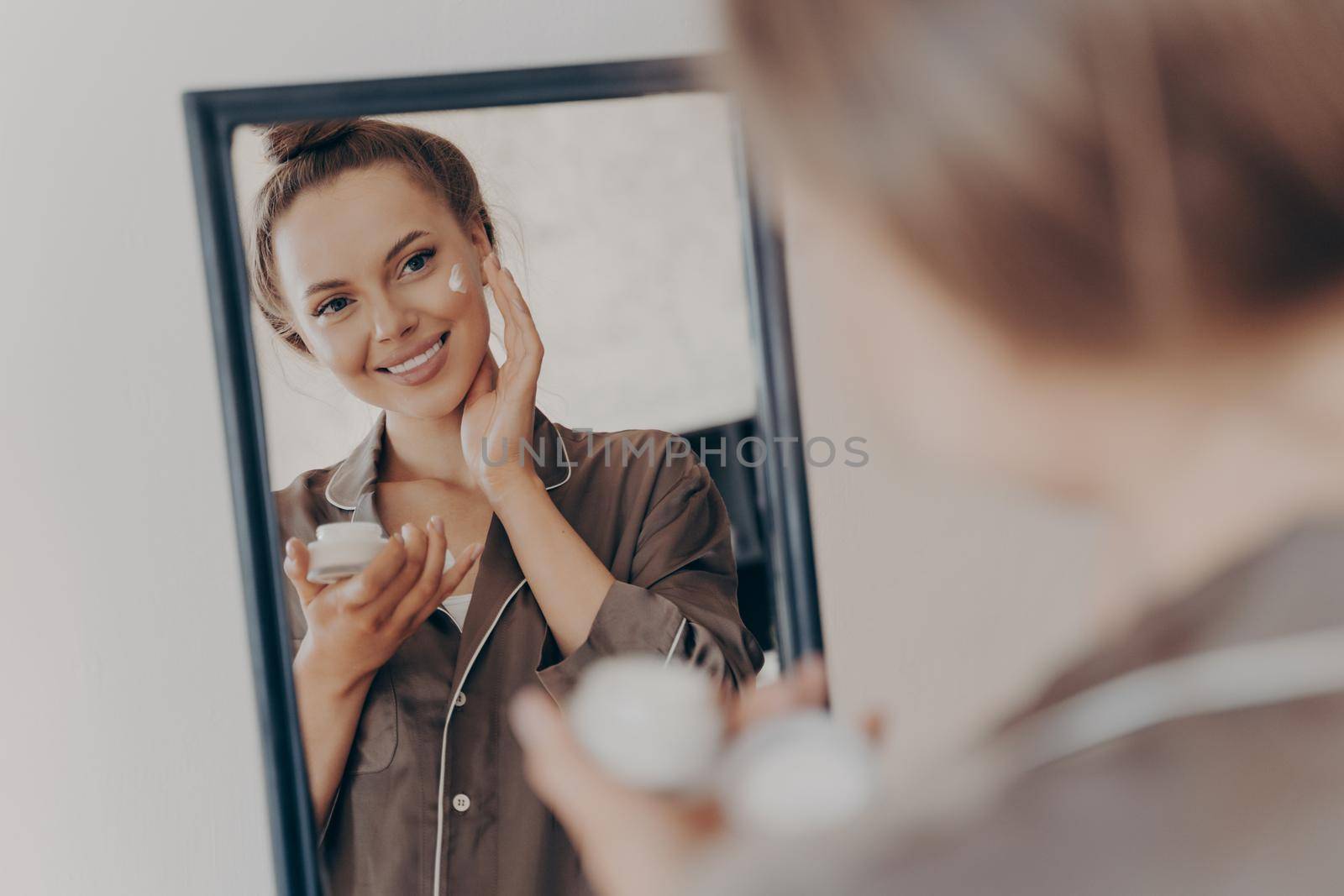 Reflection in mirror of beautiful woman in silk pajama smiling while applying face cream for deeper repair, protecting facial skin. Skincare, beauty and effective anti wrinkle treatment concept