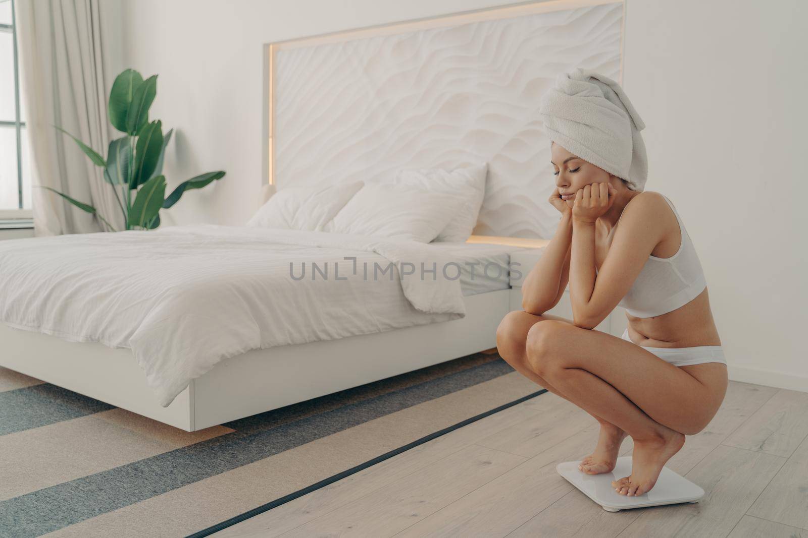 Frustrated young caucasian girl standing barefoot squatted on electronic scale in underwear after taking shower in modern bedroom next to large bed, feeling sad. Weight loss and dieting concept