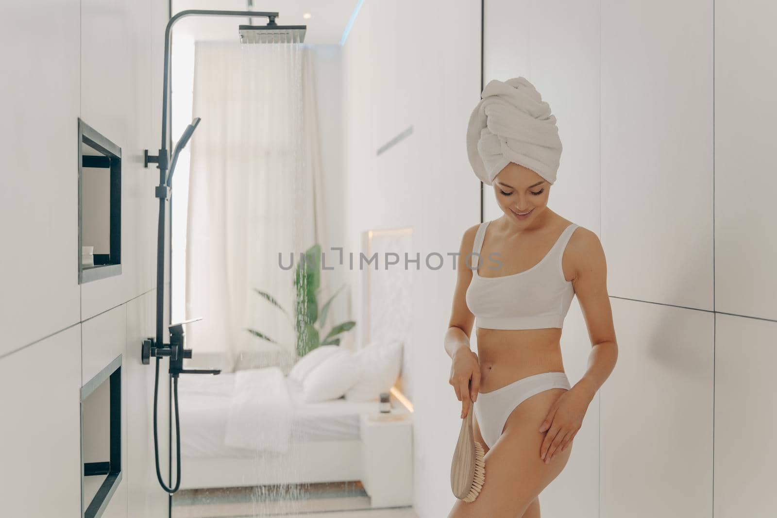 Attractive young lady in white underwear doing scrubbing massage with dry brush before morning shower in light colored bathroom on background of stylish bedroom. Women beauty and hygiene concept