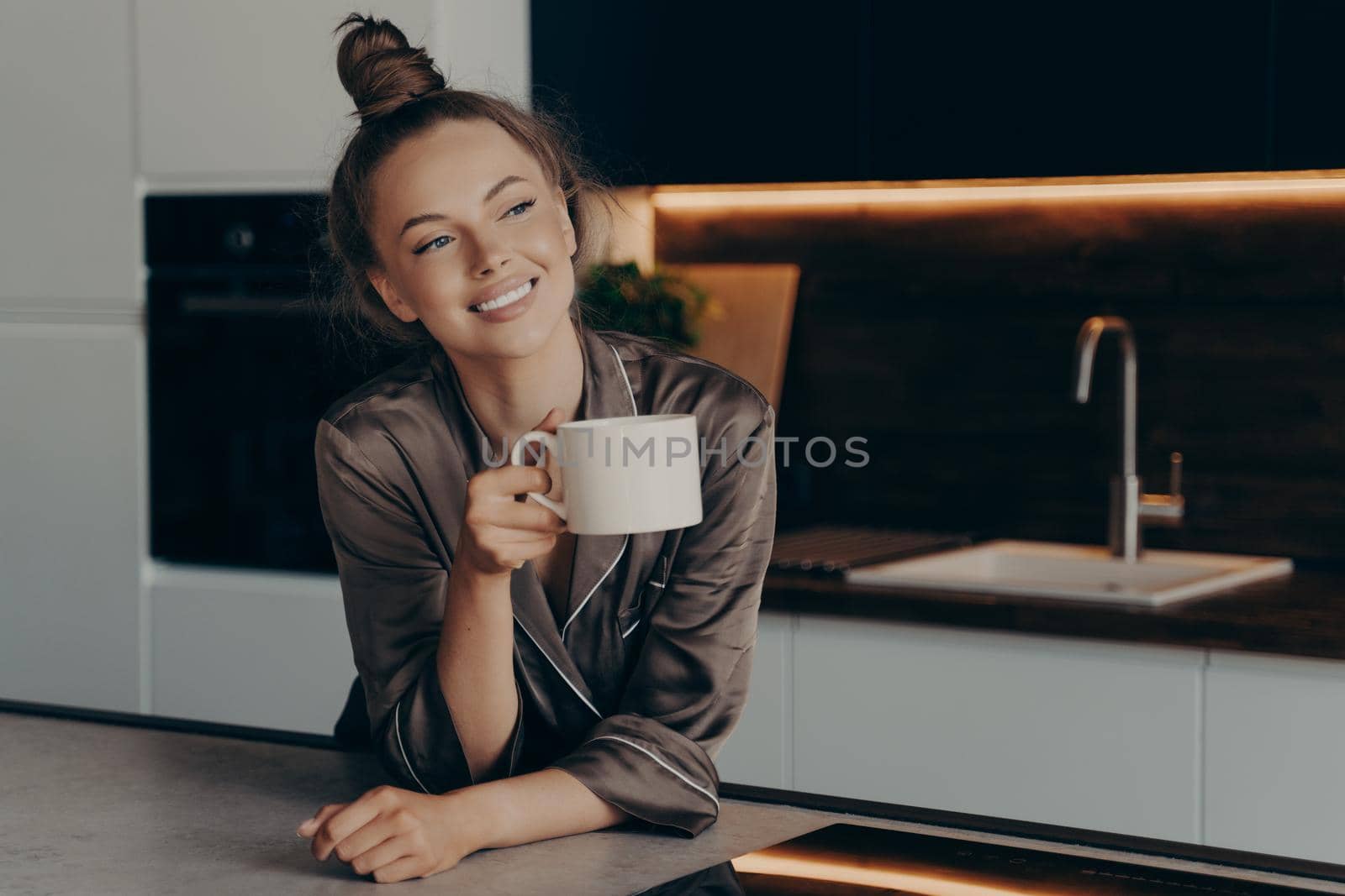 Happy smiling young woman in brown satin pajama enjoying aromatic morning coffee in kitchen by vkstock