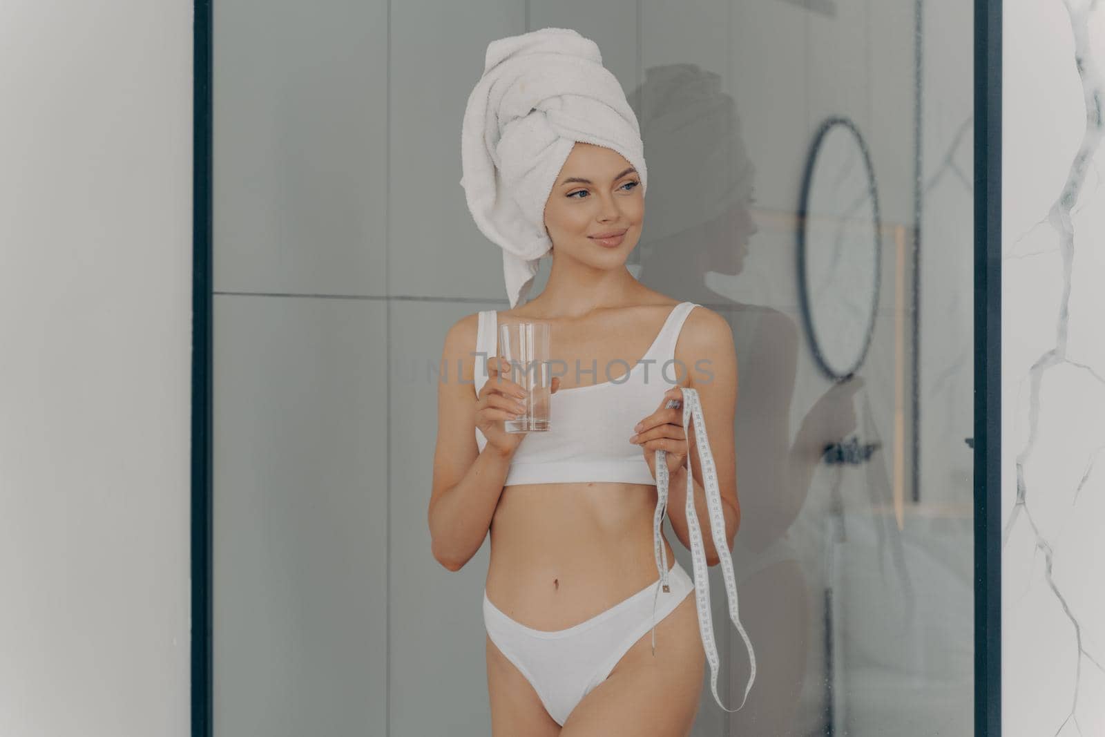 Healthy lifestyle. Fit beautiful young girl posing in underwear in bathroom after taking shower with glass of fresh water and measuring tape, happy to start early morning routine