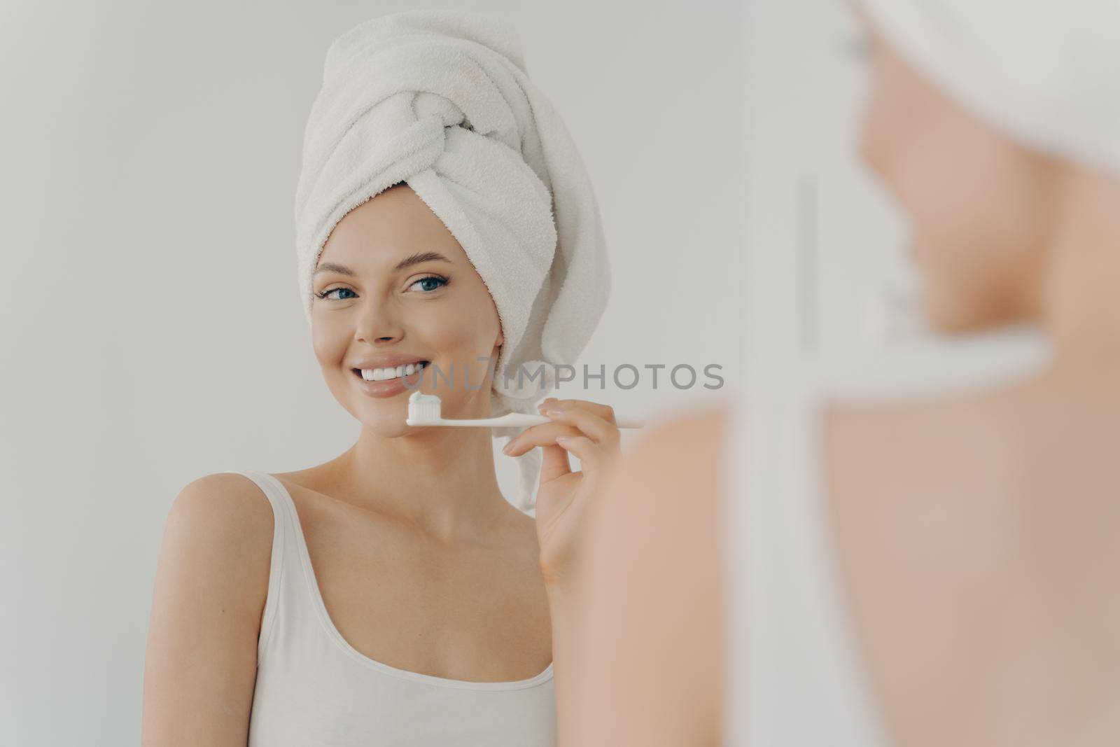 Beautiful young woman with healthy perfect smile brushing teeth and looking in mirror, attractive young female wearing white bath towel on head standing in bathroom at home. Oral hygiene concept