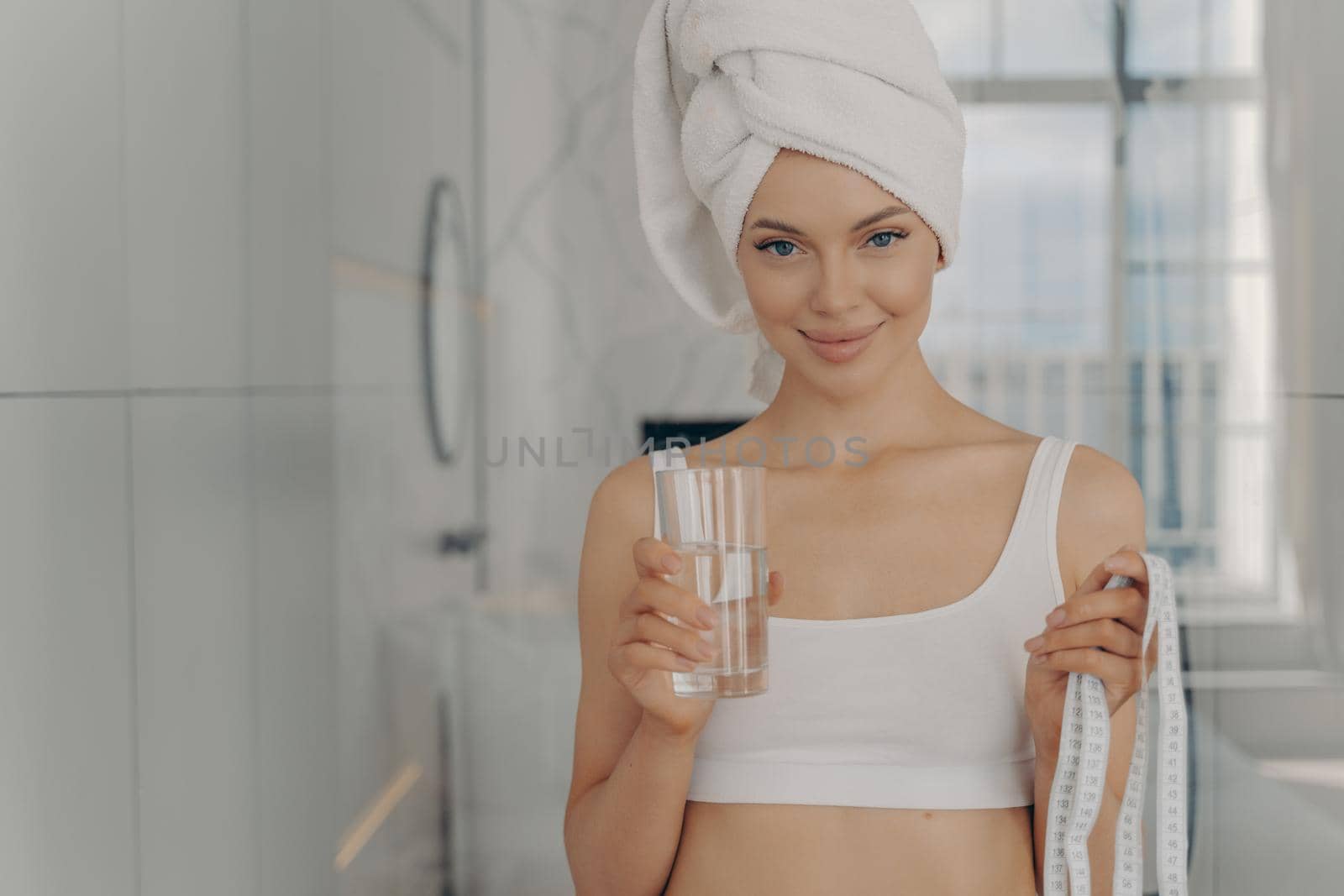 Portrait of beautiful young female with fresh glowing skin, fit figure and smiling face, holding glass of mineral water in one hand and measure tape in other and smiling at camera. Healthy lifestyle