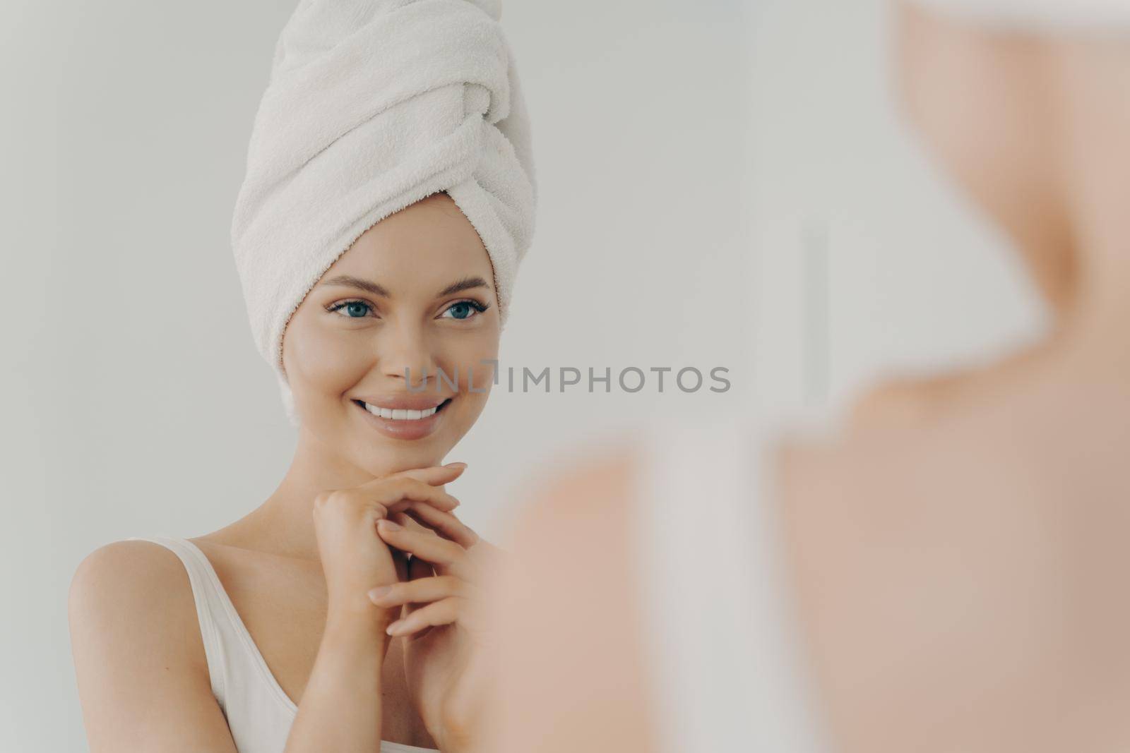 Positive healthy young woman smiling while applying facial cream reflecting in mirror, happy attractive lady putting moisturizing nourishing cream during morning beauty routine in bathroom