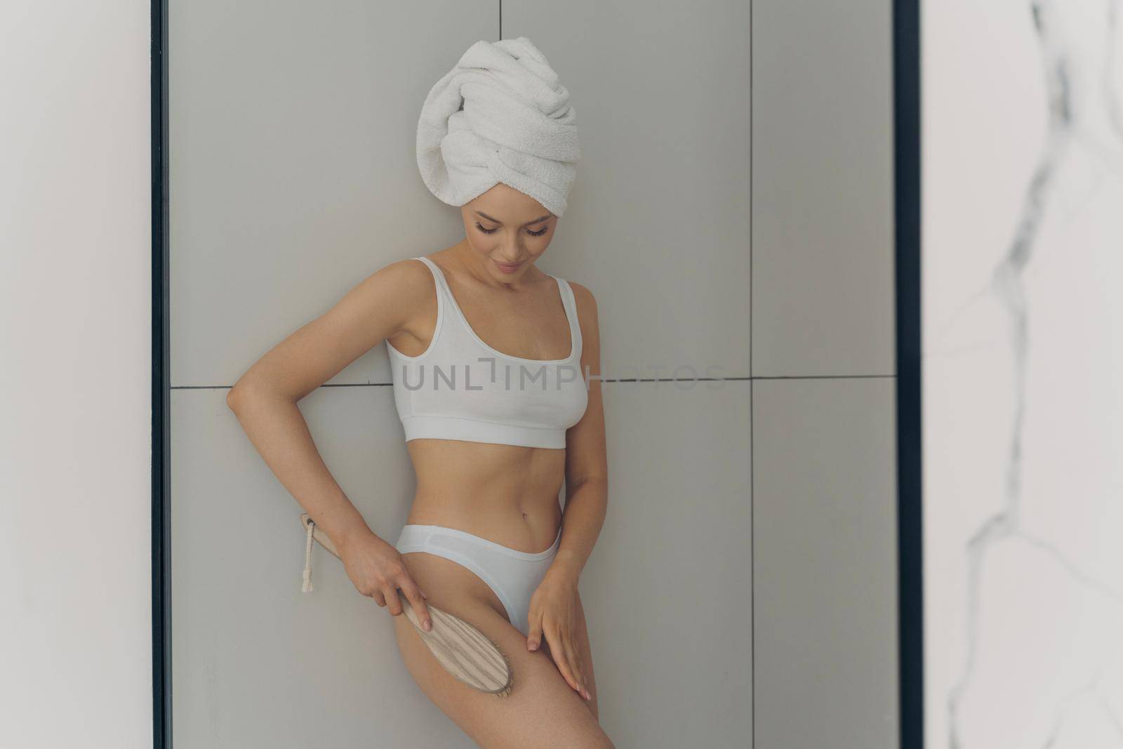 Photo of young beautiful woman doing body massage with dry brush in bathroom after morning shower, wears classic lingerie with head wrapped in towel, isolated on white marble wall background
