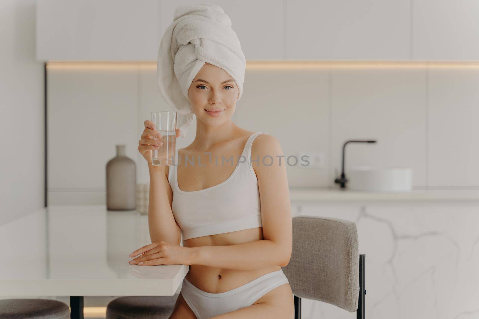 Good morning. Portrait of young slim female model posing with glass of water and smiling at camera, sitting in kitchen in white classic underwear and bath towel wrapped on head. Healthcare concept