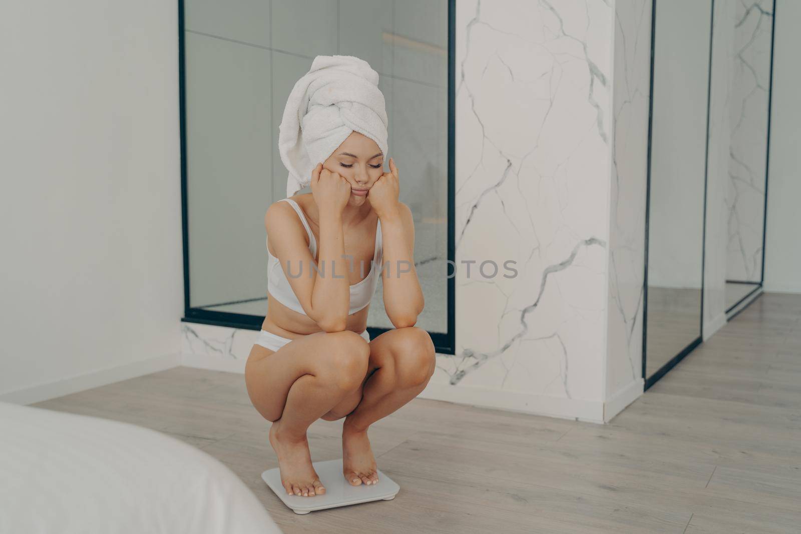 Young unhappy slender caucasian girl squatting barefoot on electronic scales with sad face on hands in stylish bedroom interior, female could not lose weight during dieting