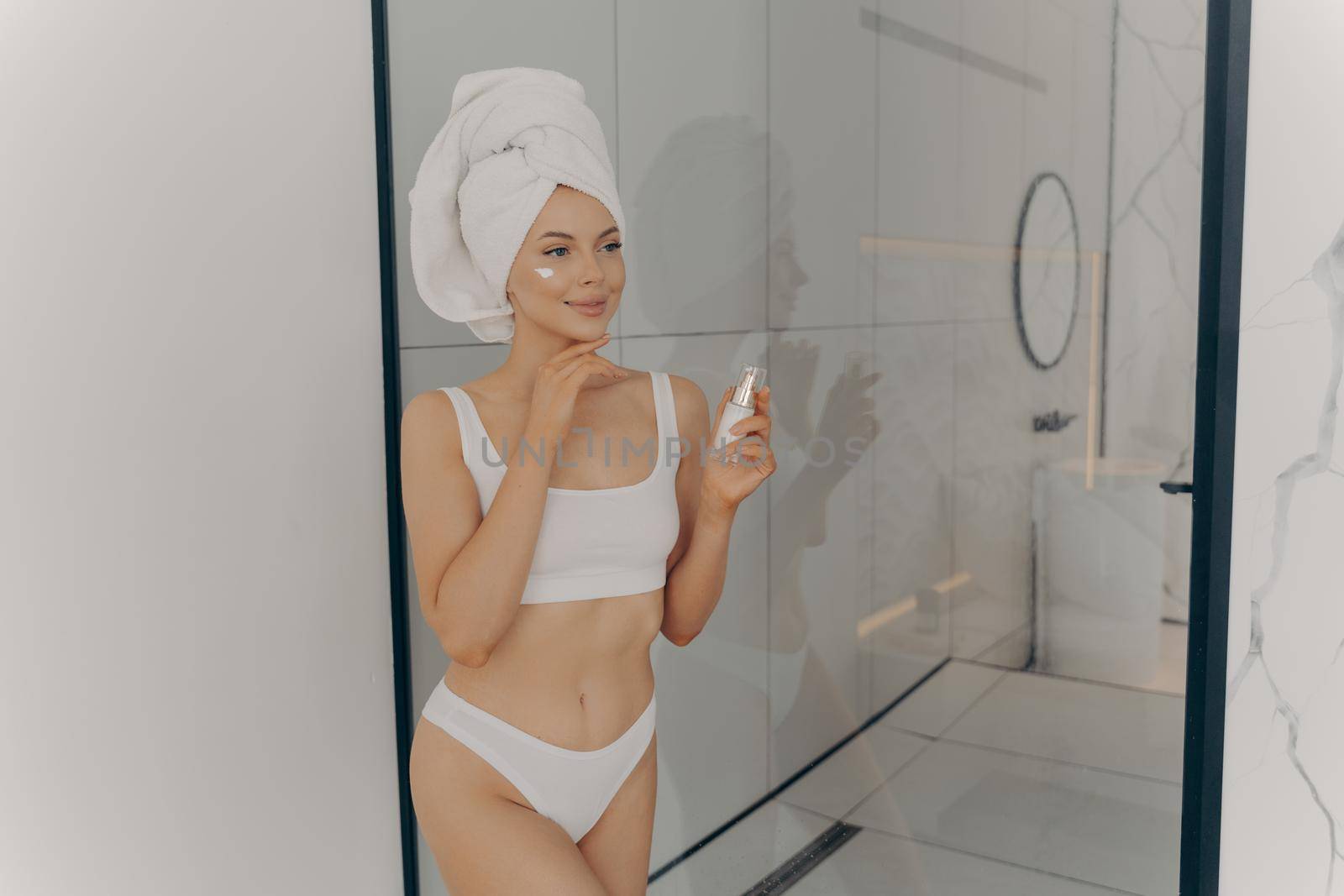 Attractive young adult woman applying facial cream after morning shower by vkstock