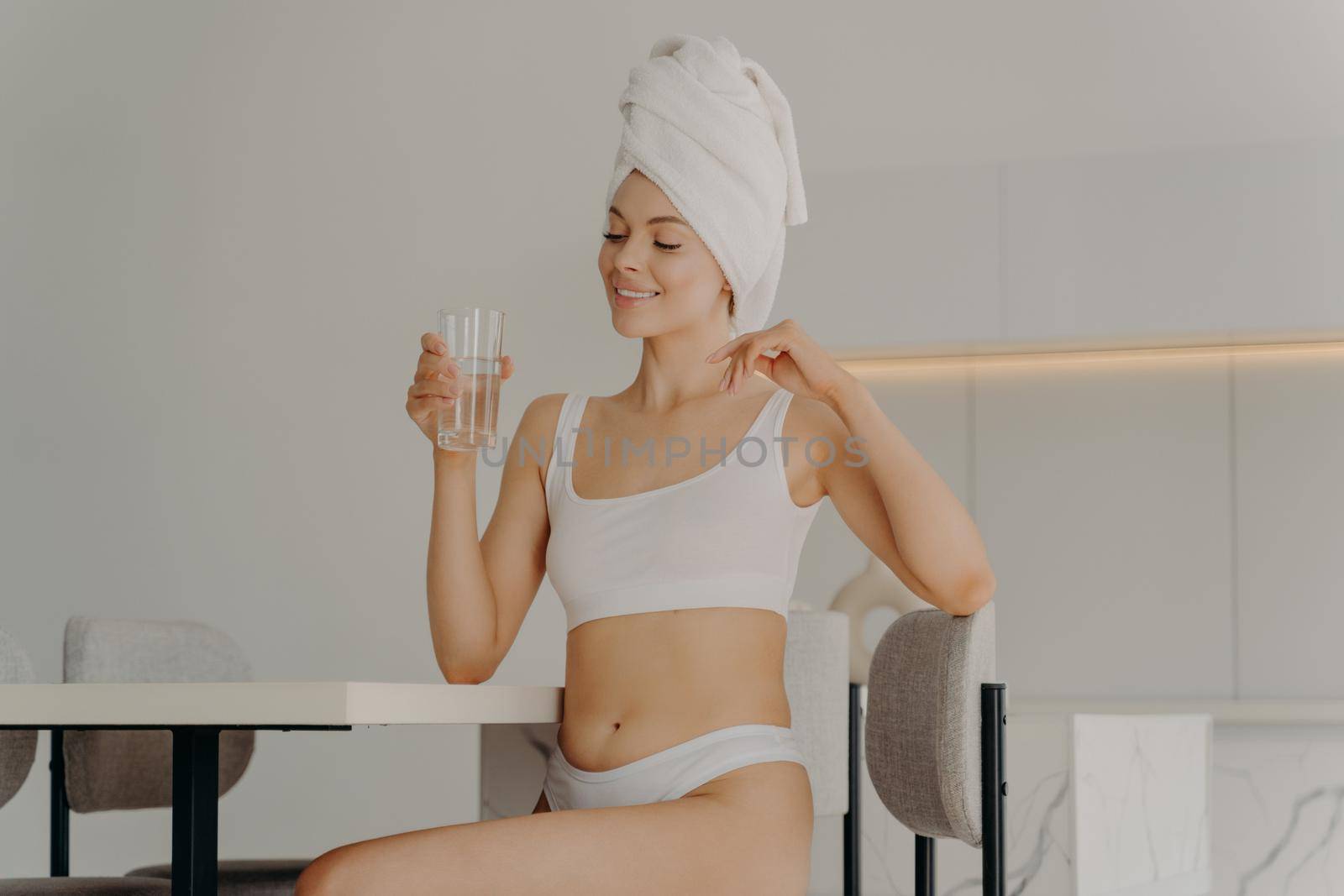 Happy slim young woman sitting on chair in kitchen with glass of pure water in her hand, relaxing after morning refreshing shower and starting new day with healthy habits. Dieting concept