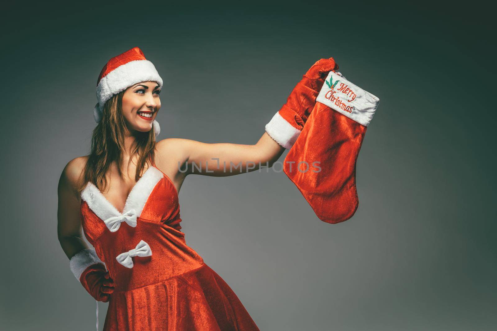 Beautiful young smiling woman in Santa Claus costume thinking and holding red christmas stocking.