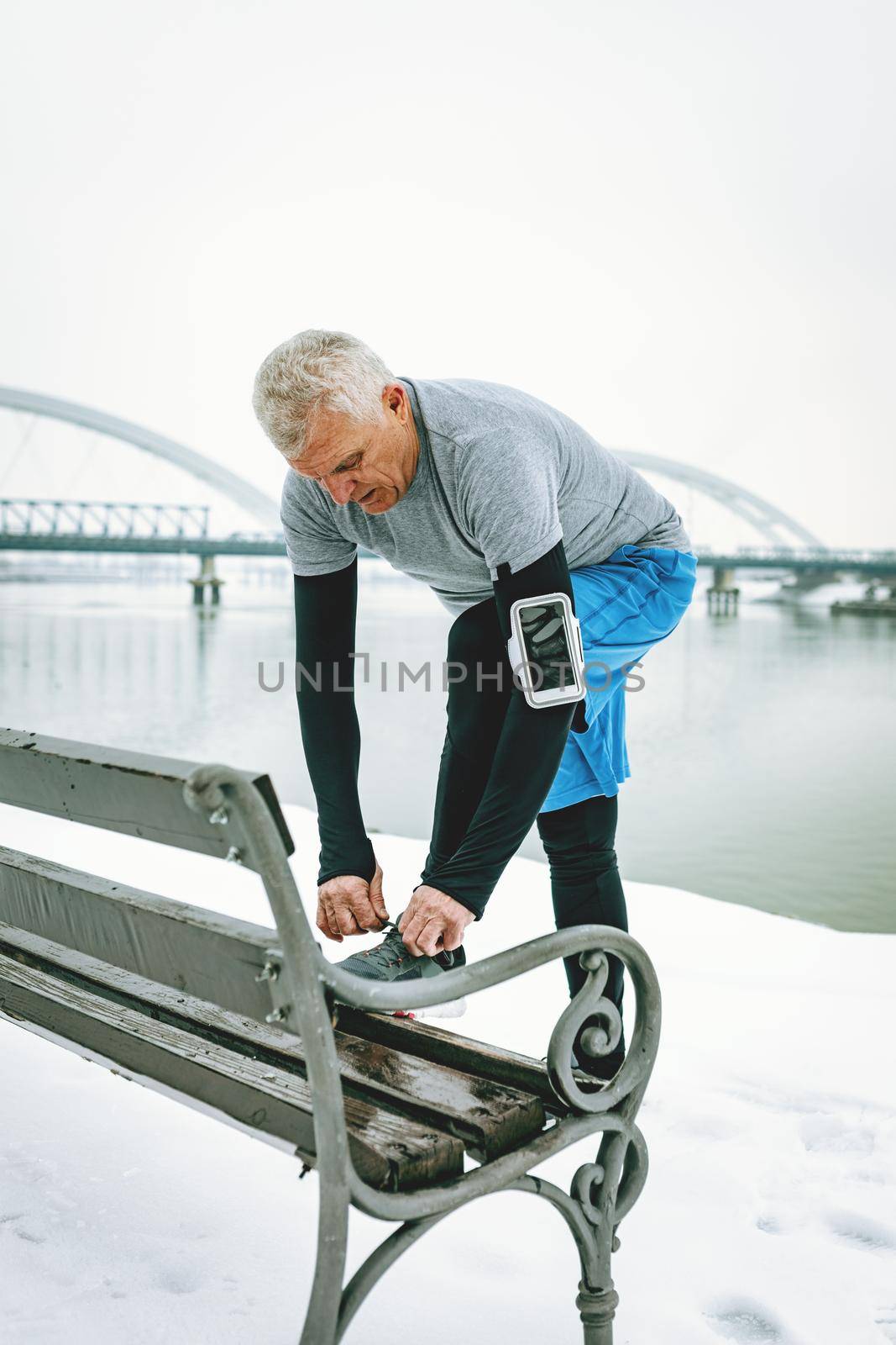 Active senior tying running shoes, and making pause during stretching and doing exercises by the river during the winter training outside in.