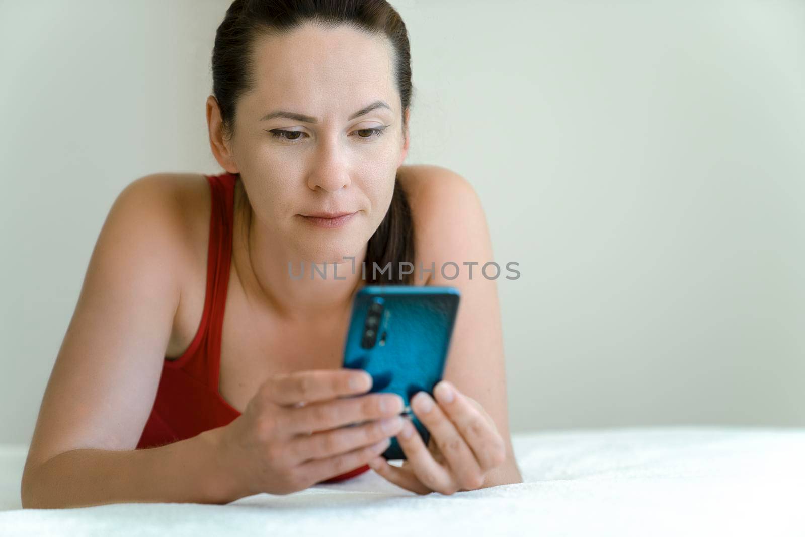 Girl holds smartphone in her hands and communicates via video communication with neutral emotion on her face lying on bed at home.