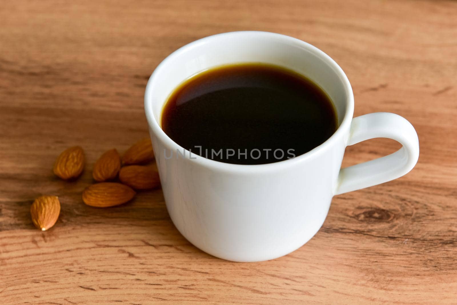 White cup of strong black coffee with almonds lying nearby on a wooden surface close-up.