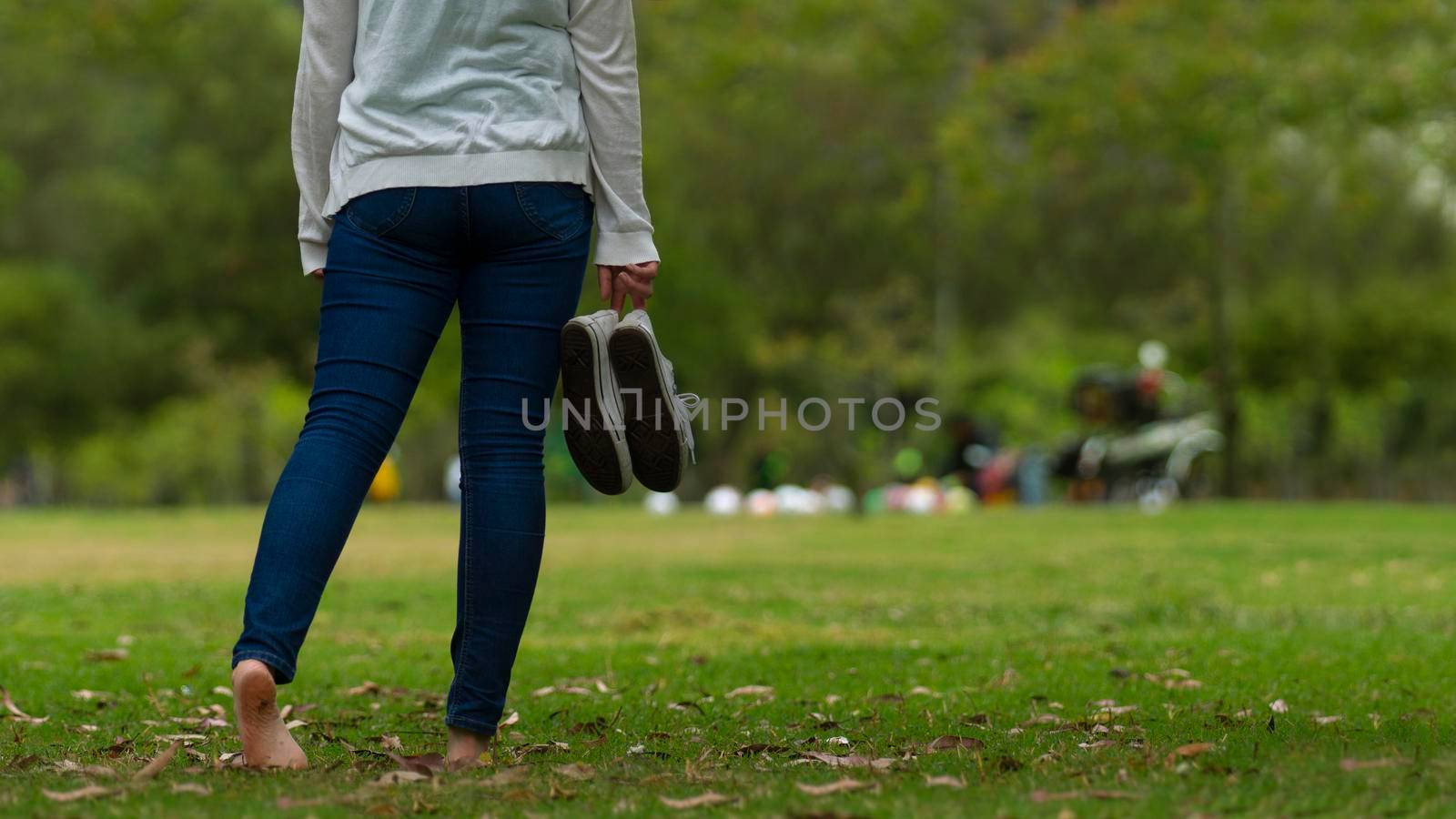 Closeup to young woman dressed in blue pants walking barefoot on the grass in the park, holding her sports shoes in her right hand with green trees background