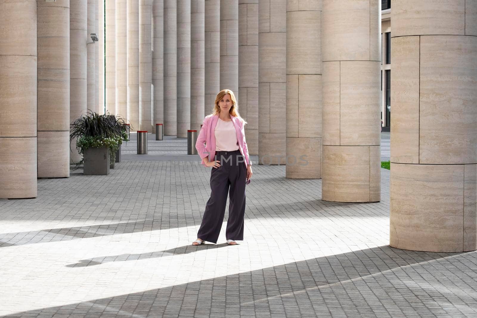 Young elegant beautiful blonde millennial with long hair in pink clothes stands in the colonnade of modern building. Full height. Selective focus.