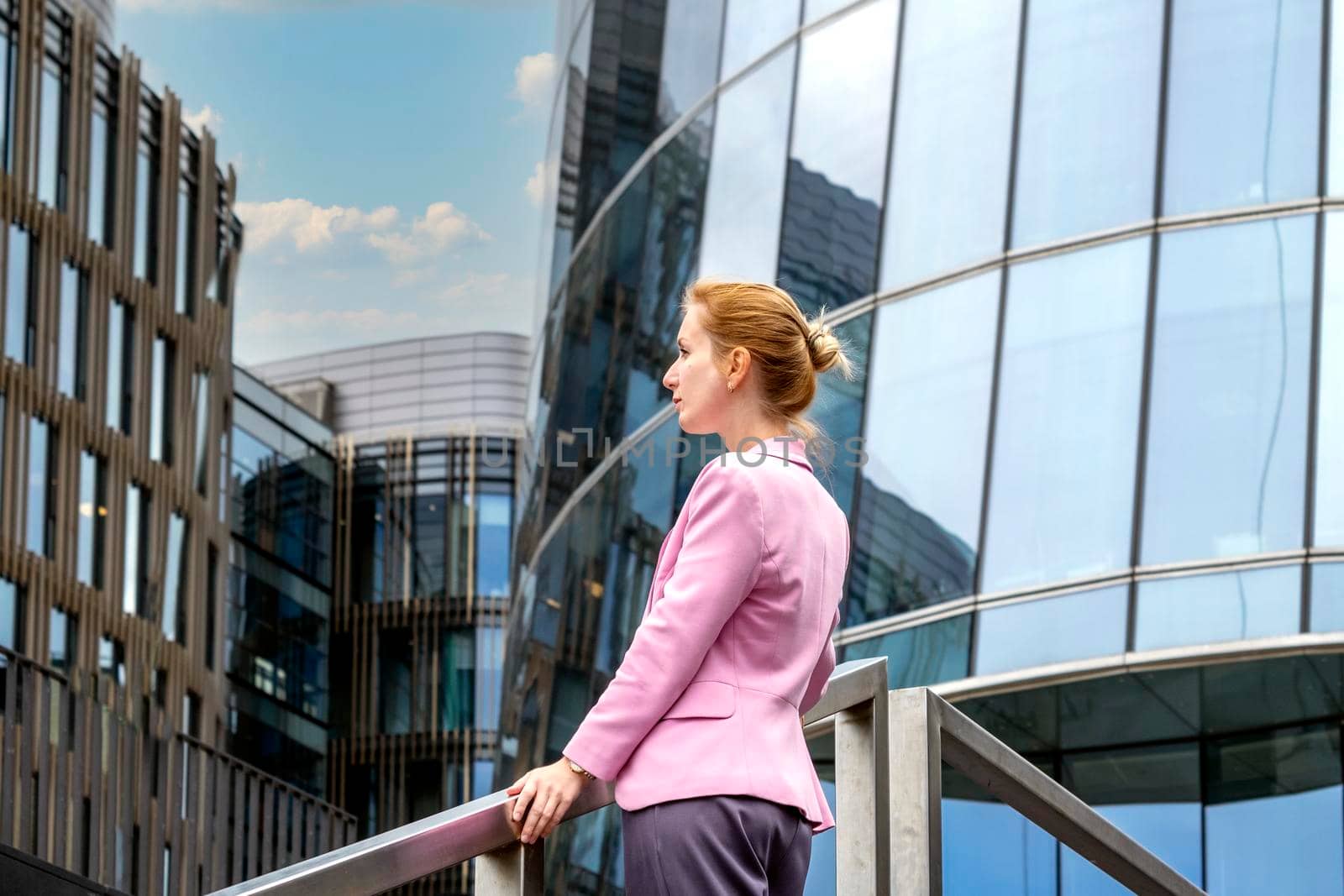 Young blonde with tailored hairstyle in pink jacket stands on steps and looks out at modern office building made of glass and steel. Profile view. Selective focus.