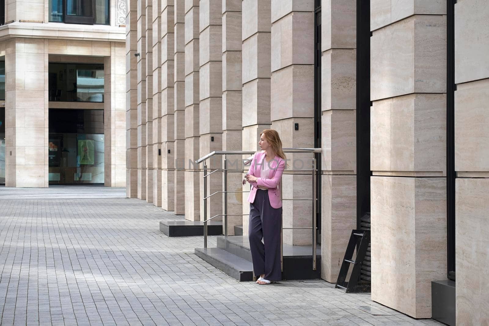 Young elegant beautiful blonde millennial with long hair in pink clothes is standing near a modern building. Full length, erase down. Selective focus.