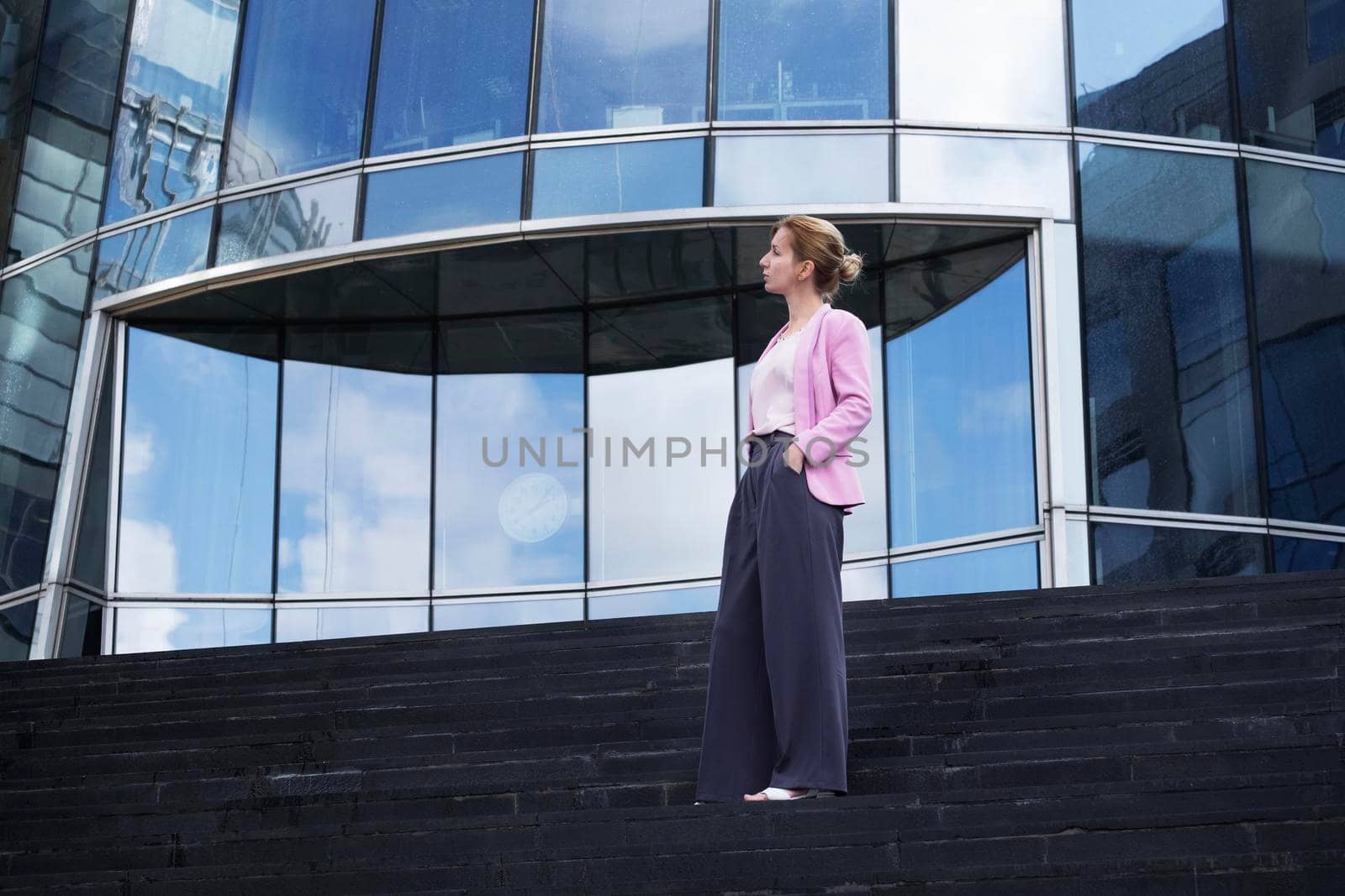 Young blonde of millennium with strict hairstyle in pink elegant clothes stands on steps near modern office building in full growth, half-turned to camera. Selective focus.